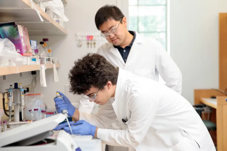 One man in white lab coat uses pipette while another supervises